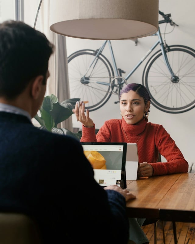 A man and a woman are having a conversation at a table over their laptops.  A bike is hanging on the wall in the background