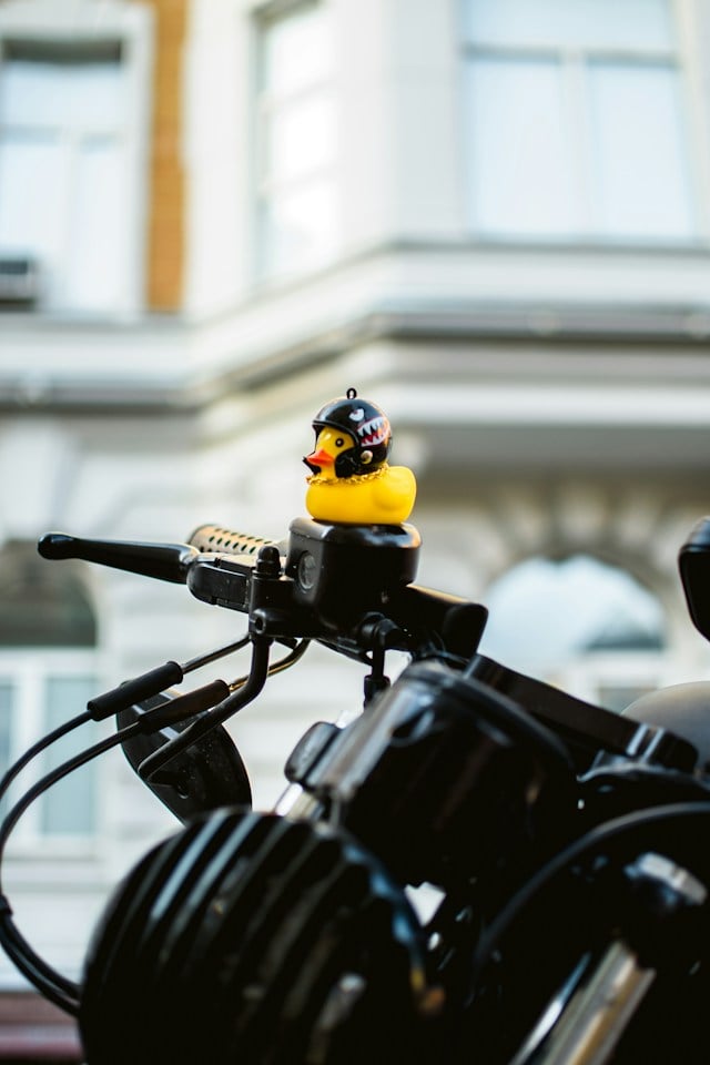 A small yellow rubber duck sitting on the handlebars of a motorcycle wearing a helmet