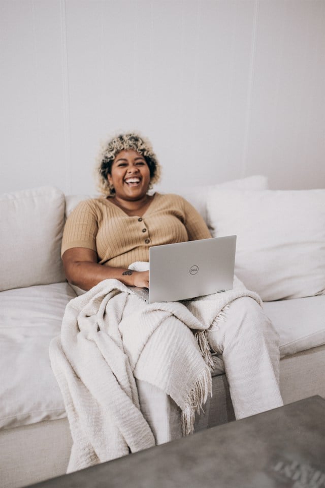 A woman sat on a white sofa laughing with a laptop on her lap
